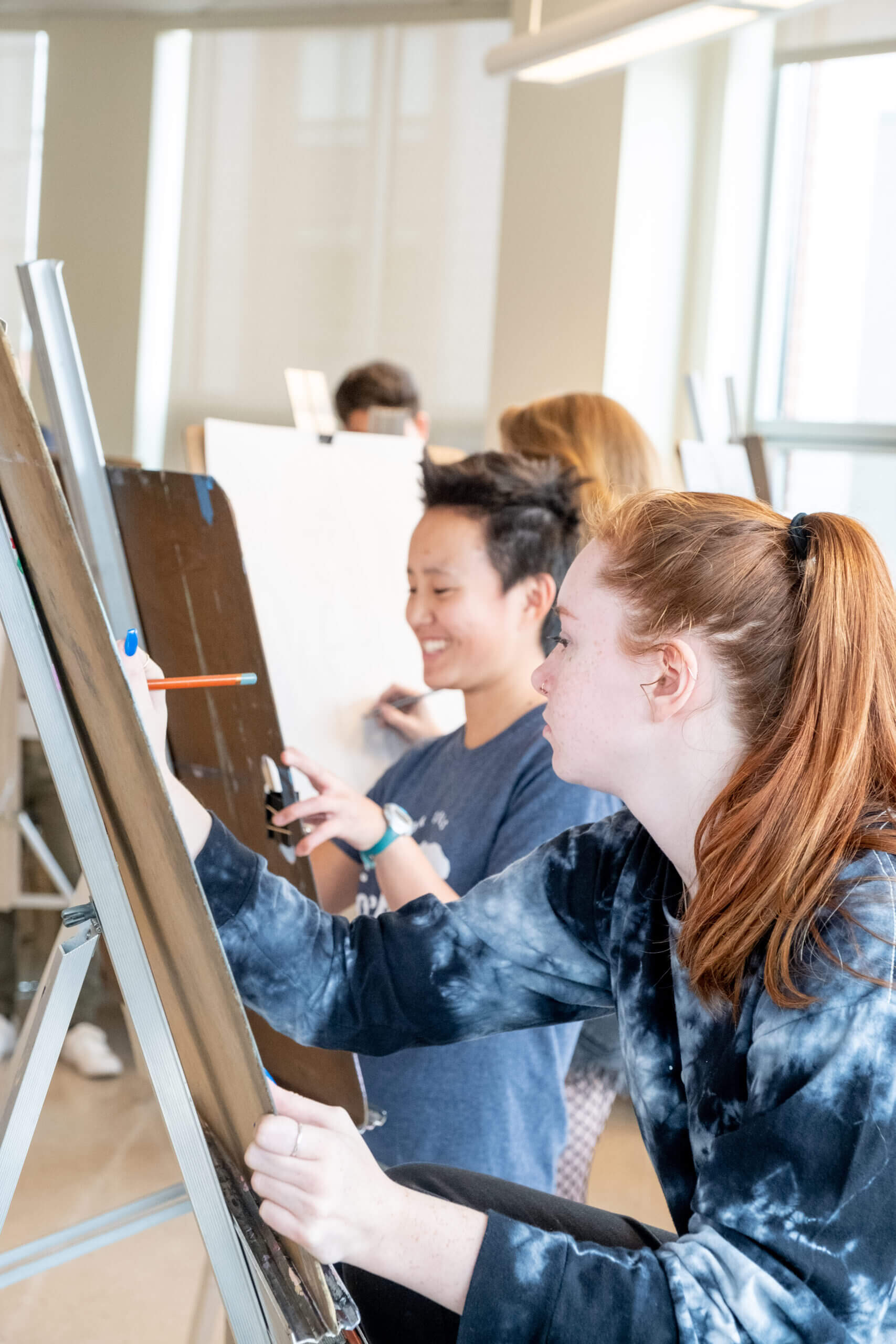 Two students painting on easels in art classroom, smiling and focused