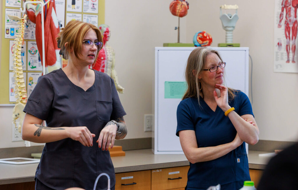 Two students listen to their instructor during a medical assisting lab.